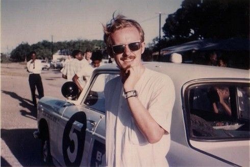 Old photo of a young white man with sunglasses and a mustache leaning against a car. He looks pretty cool.
