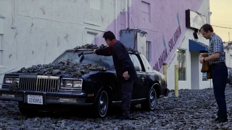 still from a 1990s film, depicting two men cleaning hundreds of dead frogs off the hood of a car
