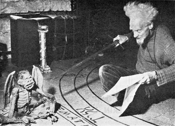 Black and white photo of a very quirky looking old man with frizzy hair, sitting in a magic circle and holding a sword. He is pointing the sword at a clay figurine of a demon as though it might attack him at any moment.