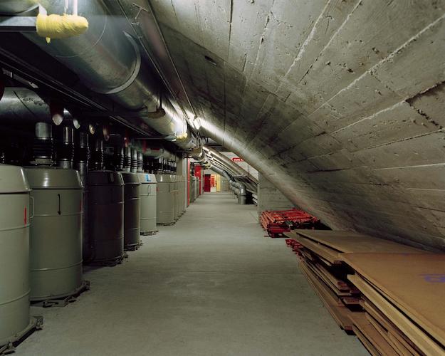 Photograph of a claustrophobic concrete tunnel lined with cardboard pallets and grey tanks. The ceiling slopes down threateningly.