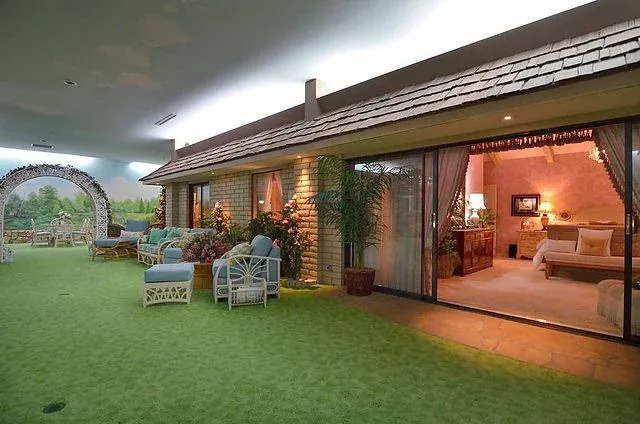 Photo of a bizarre interior designed to look like an exterior, with fake grass carpet and sky painted on the ceilings. The adjoining room is designed to look like the "inside" of a house. Everything is in a creepy, chintzy 1950s style.