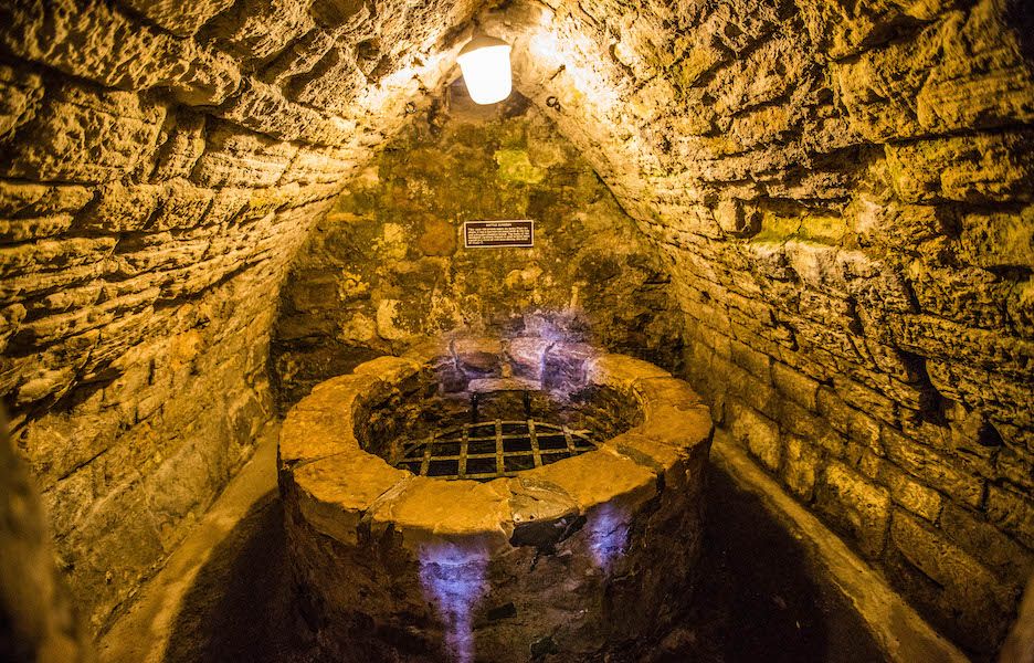 Interior of a medieval dungeon with stone walls and a well covered by an iron grate. It is lit by a modern electric lamp.