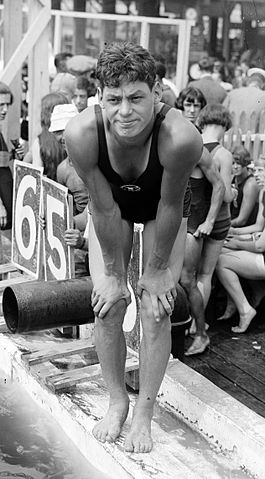 Black and white photo of a keen-looking young man in a swimmer's one piece, stretching as he presumably readies himself to swim