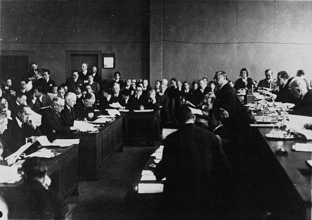 Black and white photo of a room full of suited politicians at desks, mostly white men, with a few Chinese men speaking in the centre of the frame