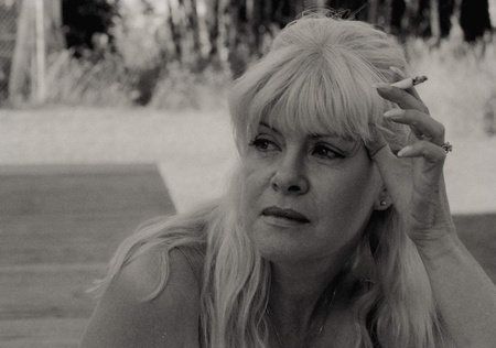 A black and white photo of a middle-aged blonde woman smoking a cigarette. She looks weathered and world-weary.