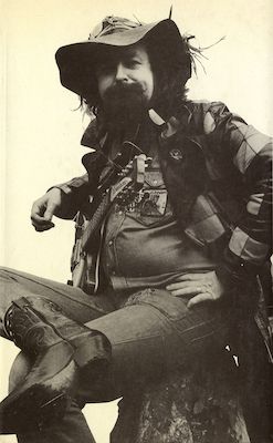 Monochrome photograph of a scruffy, bearded white man, wearing a floppy hat and cowboy boots, with a guitar across his knees. He looks like a cross between Woody Guthrie and a wizard.