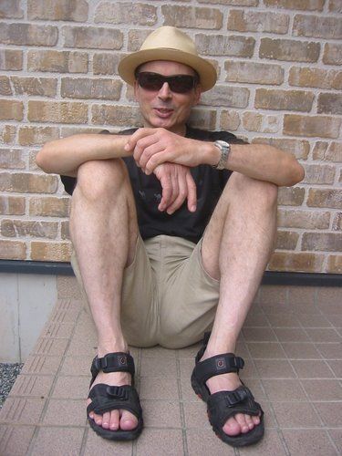 A white man sitting on the ground, leaning his back against a brick wall. He is wearing shorts, sandals, wraparound sunglasses and a beige fedora. He looks like an incredibly chill guy.