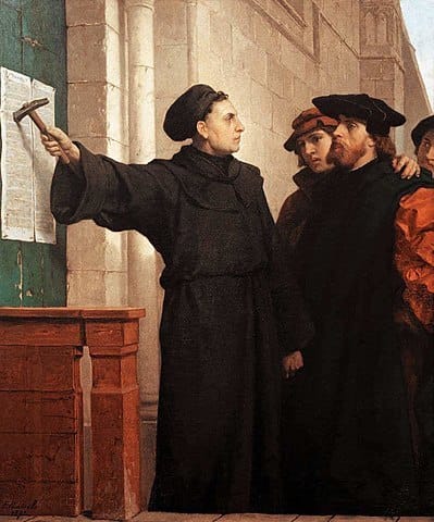 Painting of Martin Luther, in a black robe and cap, nailing a set of papers to a green door. He holds out his hammer dramatically while several townsfolk look on in consternation.