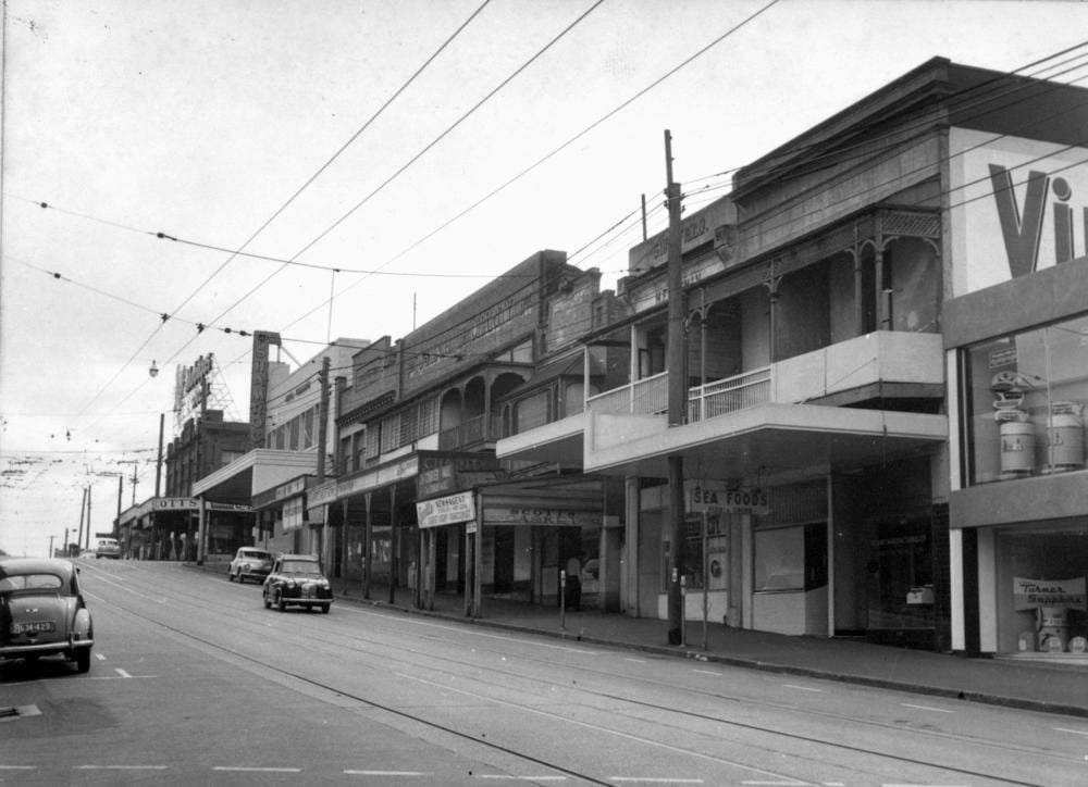 Black and white photo of an Australian city street. There are few cars on the road, no people, and the shops look rather run-down and seedy.
