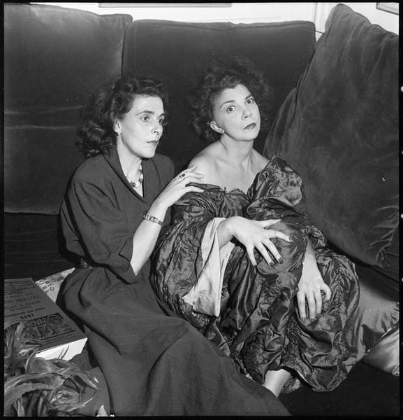 black and white photo of two women sitting on a couch, posed in an odd theatrical way, one clutching the other's shoulder