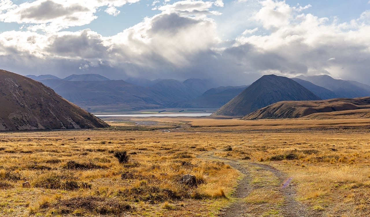 stark, cold grassland beneath a sky full of huge clouds, with slumping mountains in the distance surrounding a silver lake.