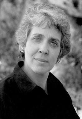 Monochrome photo of a woman with short grey hair, silver earrings and a black collared shirt. She looks... kind of wise but a bit hard-edged as well.