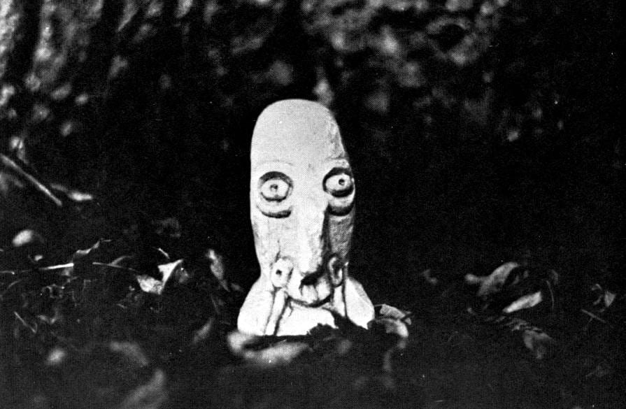 Grainy photograph of an eerie stone sculpture sitting among leaf litter on the ground